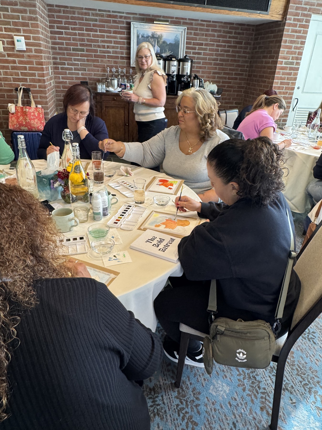 People painting colorful autumn leaves at a shared table during Jane’s watercolor gathering